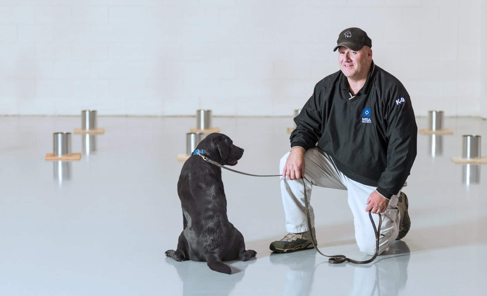 black labrador retriever and handler sitting in training facility surrounded by explosive training devices
